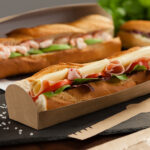 Close up of baguette tray, filled with ham, cheese, and salad baguette. Additional baguettes in background.