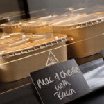 Stagione pots with clear lids, on black shelf in hot hold cabinet. Food contents description label stating 'mac and cheese with bacon'.