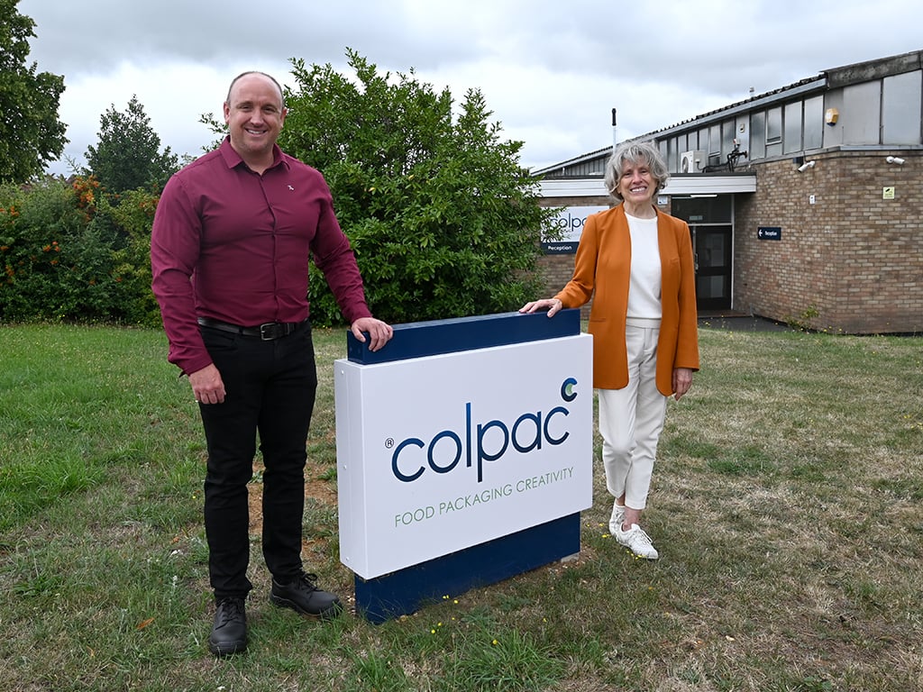 Man and woman stood either side of Colpac monolith sign outside Colpac office building. Man wearing red shirt and black jeans, woman wearing orange jacket with white top and trousers.