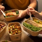 Close up of paperboard Stagione pots on a table, with two people holding forks within the pots. Pots filled with grilled fish, cous cous and vegetables, and two other pots filled with pasta and ribs on table.