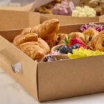 Close-up of a paperboard platter trays, filled with croissants, pain au chocolat, fresh berries, and decorative flowers on a marble surface.