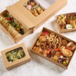 Overhead view of assorted platter trays containing sandwiches, wraps, pastries, savoury snacks, mini quiches, frittata slices, and fresh garnishes, with some platters featuring clear windowed sleeves and lids.