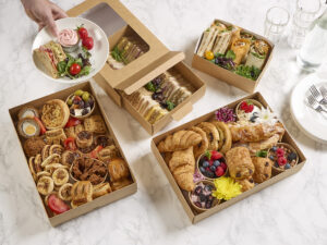Overhead view of a variety of paperboard food platters, filled with sandwiches, wraps, pastries, cakes, savoury snacks, and fresh fruit, with a hand serving a plate containing a cupcake, strawberries, sandwiches, and salad.