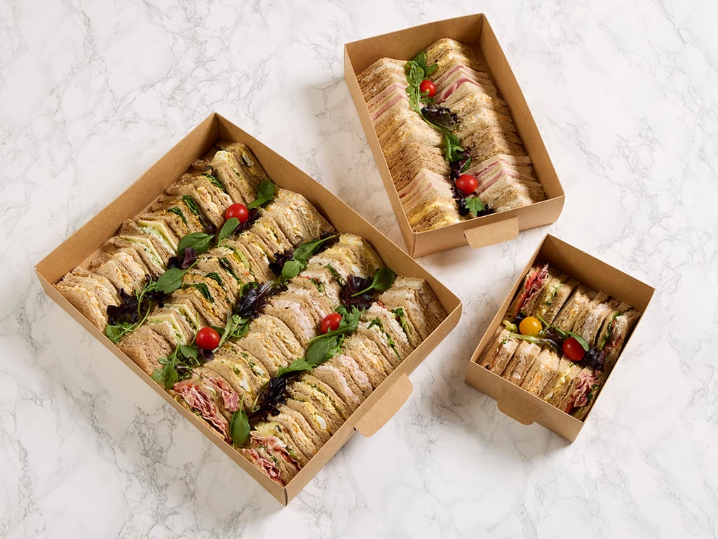 Overhead view of three paperboard food platters filled with sandwich wedges, garnished with salad leaves and cherry tomatoes, placed on a marble surface.