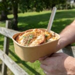 Stagione pot filled with pie, mashed potatoes, gravy and spork. Pot being held by person leaning against fence, field and trees in background.