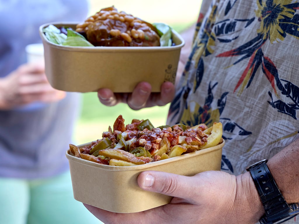 Person in floral shirt holding loaded fries and a baked potato with beans in paperboard trays.