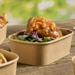Paperboard tray containing a baked potato topped with beans and salad, on a rustic wooden table.
