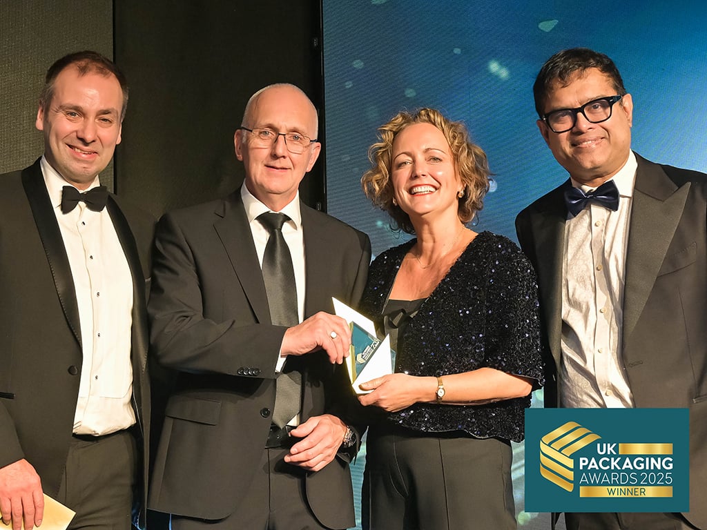 Colpac team receiving the Cartonboard Pack of the Year award at a formal ceremony, sponsored by Hubergroup. Four people in black tie formalwear stood on black stage with blue backdrop.