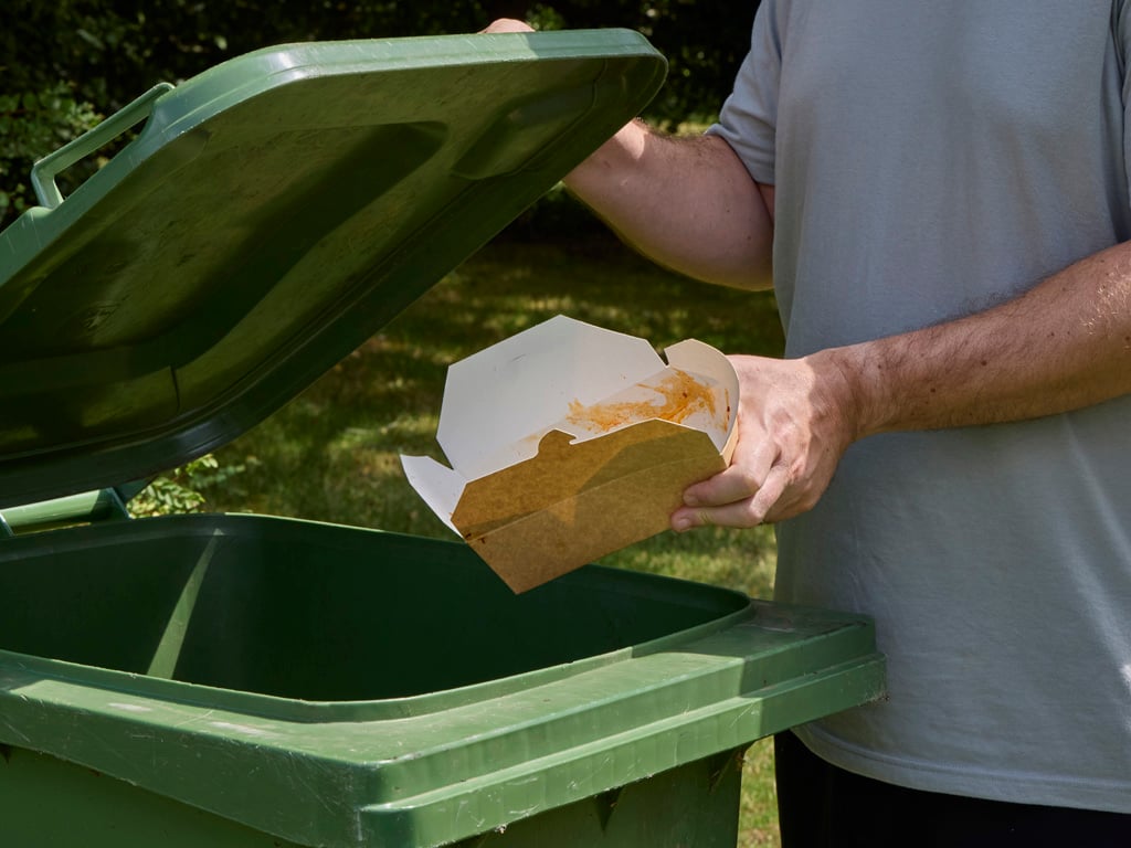 Kraft food box being put into a green bin for compostable waste. Lid being lifted by person, field in background.