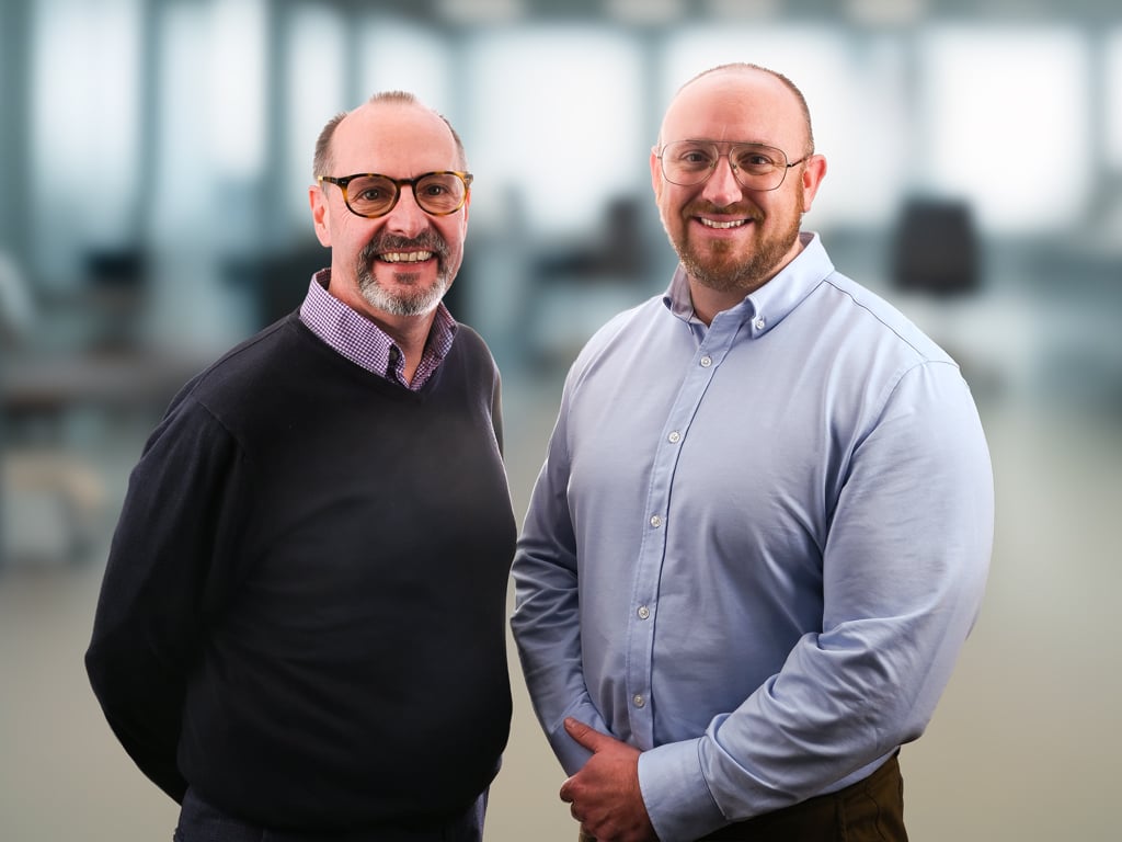 Corporate image of two men stood next to each other. Both wearing glasses and smart shirts. Out of focus office in background.