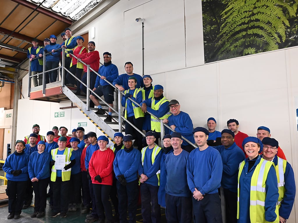 Colpac factory team stood on stairs at side of UK factory. Team stood in a group wearing red and blue sweatshirts and hats, one team member holding BRCGS certificate.