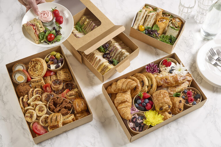 Overhead view of a variety of paperboard food platters, filled with sandwiches, wraps, pastries, cakes, savoury snacks, and fresh fruit, with a hand serving a plate containing a cupcake, strawberries, sandwiches, and salad.