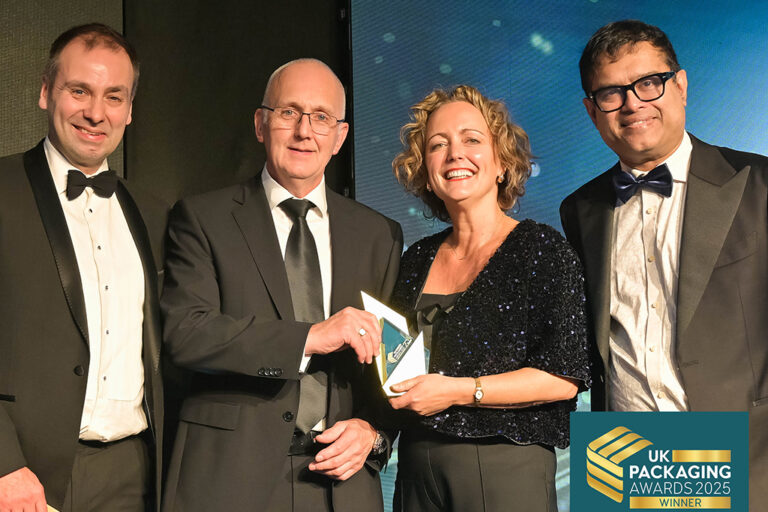 Colpac team receiving the Cartonboard Pack of the Year award at a formal ceremony, sponsored by Hubergroup. Four people in black tie formalwear stood on black stage with blue backdrop.