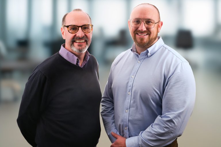Corporate image of two men stood next to each other. Both wearing glasses and smart shirts. Out of focus office in background.