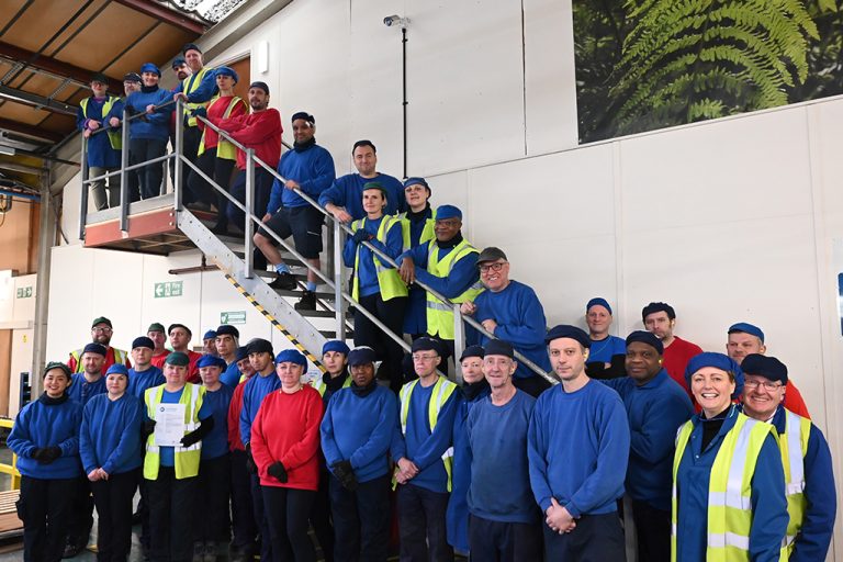 Colpac factory team stood on stairs at side of UK factory. Team stood in a group wearing red and blue sweatshirts and hats, one team member holding BRCGS certificate.